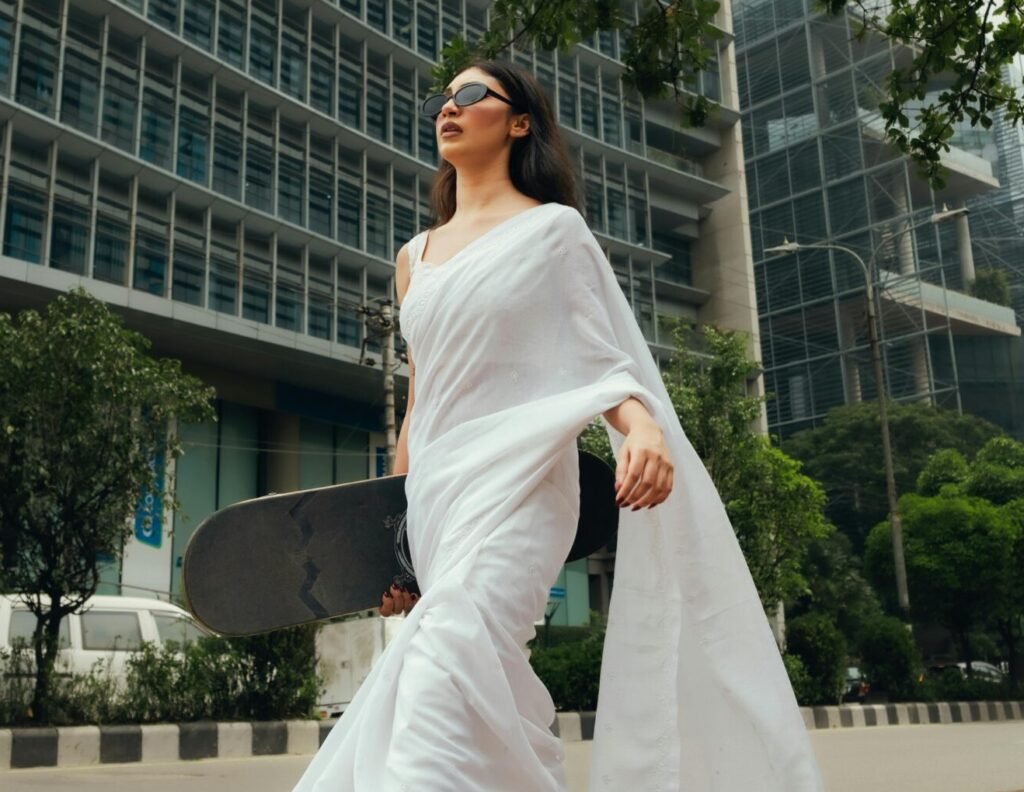 A woman in a white saree and sneakers holds a skateboard, walking through a modern urban environment.