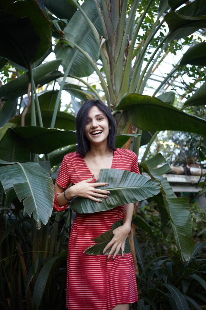 Gorgeous friendly looking dark-haired young lady in red dress with white stripes having fun in plant nursery, covering her body with two large green leaves, laughing cheerfully. Vertical shot